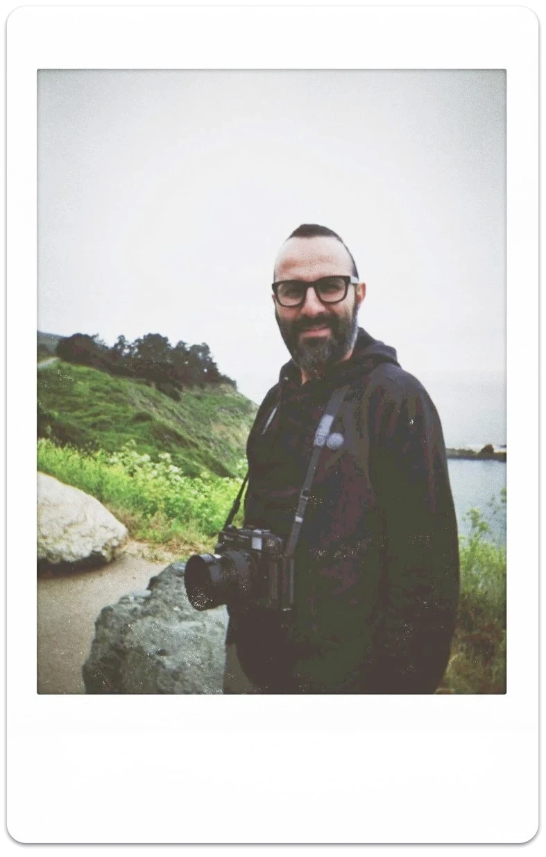 Reza smiling, with a camera around his neck, on a coastal trail